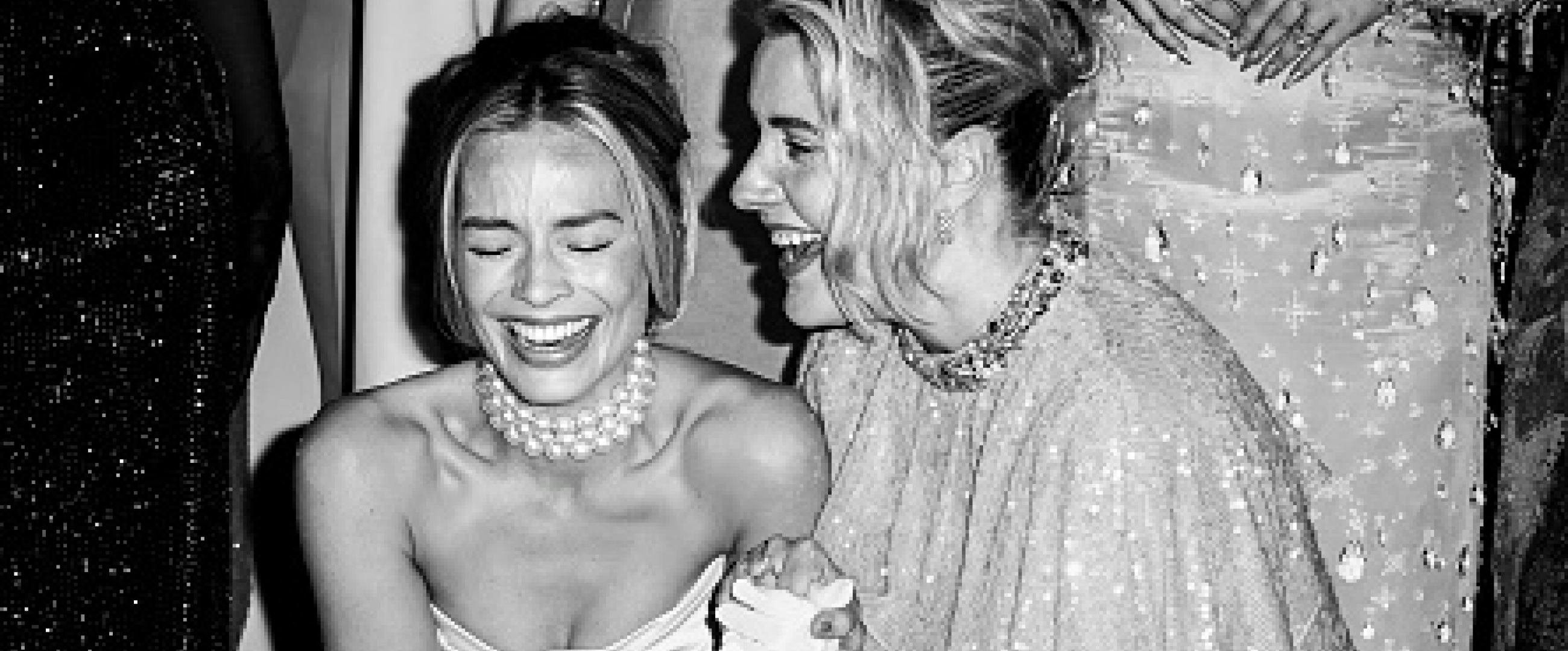 Black and white photo of producer-actress Margot Robbie and director Greta Gerwig laughing together and clutching one another's hand during a red carpet publicity event for the "Barbie" movie